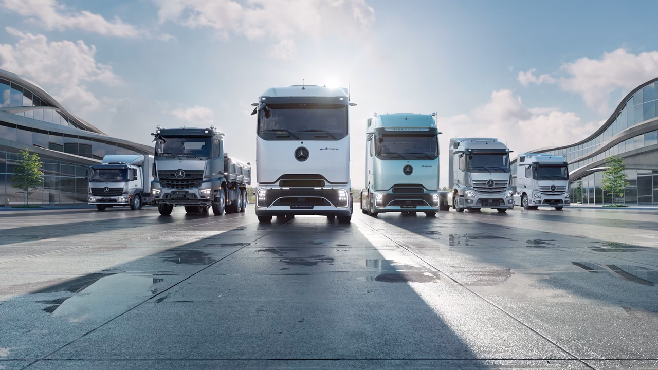 Mehrere Mercedes-Benz Lastkraftwagen stehen nebeneinander auf einem nassen Parkplatz vor modernen Gebäuden.