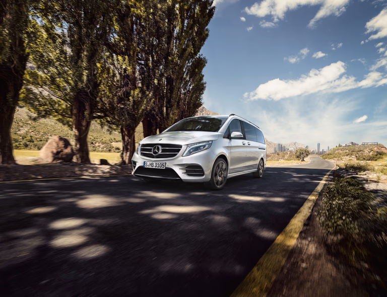 Silberner Mercedes-Benz Minivan auf Baum gesäumter Straße unter blauem Himmel mit Stadtsilhouette im Hintergrund.