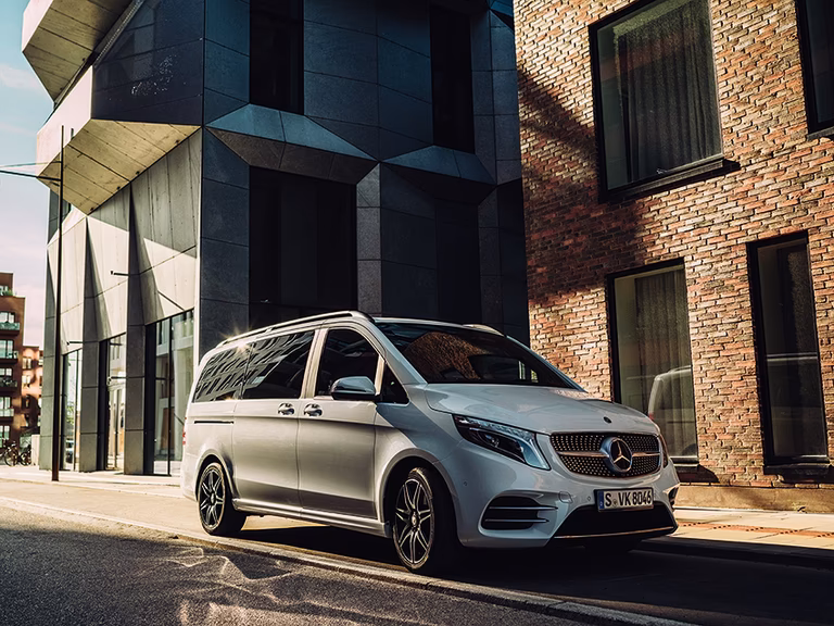 Silberner Mercedes-Benz Luxus-Minivan vor modernem Gebäude mit Glasfassade und Backsteinmauer in urbaner Umgebung.